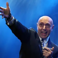 Artysta w garniturze, z podniesioną ręką, gra na harmonijce ustnej. Jego ekspresja sugeruje, że wykonuje energetyczne **shakin dudi utwory**.