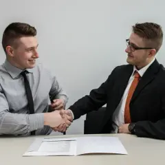 Dwóch mężczyzn w garniturach ściska sobie dłonie nad dokumentem, finalizując umowę przedwstępną sprzedaży nieruchomości.