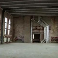 Wnętrze starej łaźni z ceglanymi ścianami, łukowymi oknami i metalowymi rurami biegnącymi po suficie i ścianach.