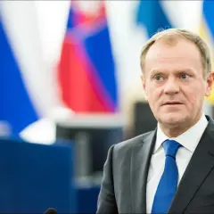 Donald Tusk w garniturze i niebieskim krawacie na tle flag.