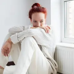 Ragazza con capelli rossi seduta vicino a una finestra, avvolta in un maglione bianco. Potrebbe riflettere su come l'olanzapina a cosa serve per affrontare momenti difficili.