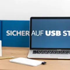 Laptop mit "Sicher auf USB Stick" auf dem Bildschirm. Ein weißer USB-Stick und eine externe Festplatte liegen auf dem Schreibtisch.