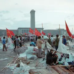 Ludzie na placu Tiananmen, czerwone flagi powiewają, budynki w tle, chaos i prowizoryczne namioty.