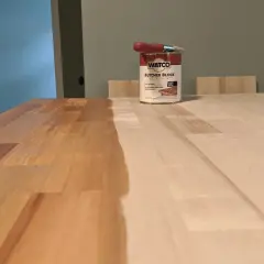 A can of Watco Butcher Block Oil & Finish sits on a wooden surface, with one half of the butcher block already stained and the other half awaiting treatment.