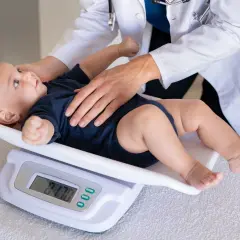Lekarz waży niemowlę na wadze cyfrowej. Wynik 7.70 kg sugeruje, ile waży 10 miesięczne niemowlę.