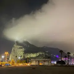 Nocny widok na Gibraltar, z chmurami otulającymi skałę. W oddali widać oświetlone miasto i palmy.