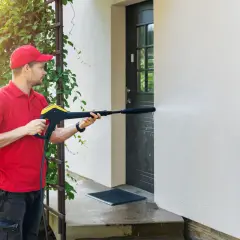 Mężczyzna w czerwonej koszulce i czapce czyści elewację domu myjką ciśnieniową. Woda spływa po ścianie, usuwając brud.