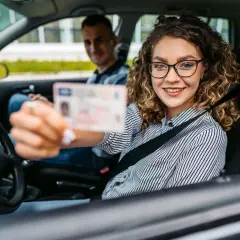Uśmiechnięta młoda kobieta w okularach pokazuje prawo jazdy, rozpoczynając swój okres próbny. Za nią siedzi instruktor.