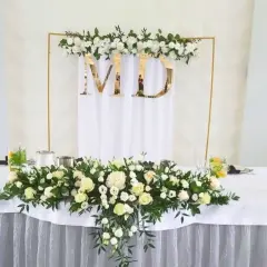 Elegancka dekoracja stołu weselnego z białymi różami i zielenią. Złote litery "MD" na tle białej tkaniny. W tle złota konstrukcja z kwiatami.