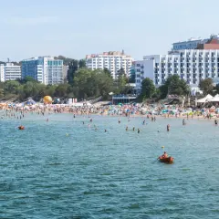 Tłum ludzi na plaży w Międzyzdrojach, kąpielisko pełne plażowiczów i łódek. W tle hotele i drzewa. Międzyzdroje atrakcje w pełnym słońcu.