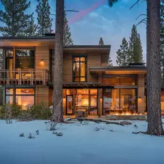 Cozy modern mountain homes with warm interior lights and a roaring fire pit, nestled amongst snow-covered trees.