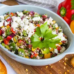 Bunter Paprikasalat mit Feta, garniert mit Petersilie, serviert in einer hellblauen Schüssel auf einem Holzbrett. Daneben liegen geröstete Brotscheiben.