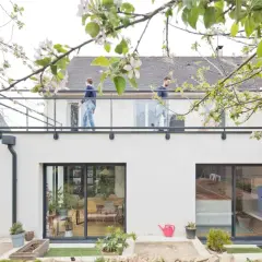 Surélévation maison 50m2 : deux personnes sur un balcon moderne, sous des branches fleuries. Le prix de ce projet est attractif.