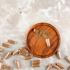 Cápsulas de maca esparcidas sobre un fondo de mármol, con un cuenco de madera que contiene polvo de maca y una cápsula abierta.