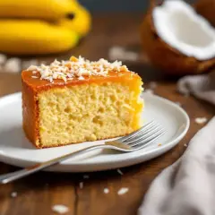 Tranche de moelleux gâteau banane sans lait, garnie de noix de coco râpée et d'un glaçage doré. Des bananes et de la noix de coco sont visibles en arrière-plan.