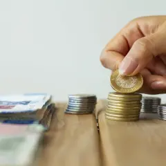 Dłoń kładzie monetę na stosie, obok leżą banknoty. To symboliczne przedstawienie oszczędności i potencjalnych korzyści z ulgi podatkowej do 26 lat.