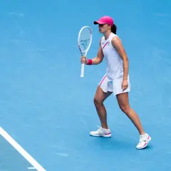 Iga Świątek w akcji, w białej sukience i różowej czapce, gotowa do uderzenia. Jej buty sportowe idealnie pasują do stroju.
