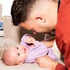 Ein Vater spielt liebevoll mit seinem 4. sprung baby auf einer Couch. Das Baby trägt einen lila Pullover und lächelt.