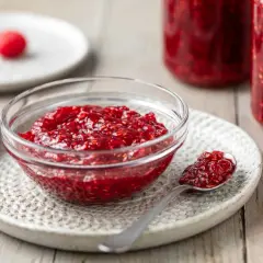 Una ciotola di vetro piena di marmellata di lamponi, con un cucchiaino e lamponi freschi sullo sfondo.