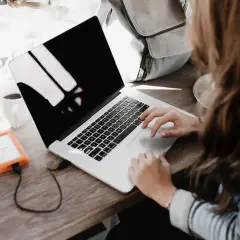 Une personne rédige une lettre de motivation française sur son ordinateur portable, avec une tasse de café et un disque dur externe à proximité.