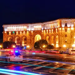 Nocna panorama placu w Erywaniu, największym mieście Armenii. Oświetlony budynek z arkadami, na ulicy widać ślady świateł samochodów i radiowóz.