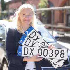 Kobieta trzyma tablice rejestracyjne z numerem DX 00398. Ułatwia to sd rejestracja.
