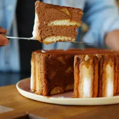 Une part de charlotte tiramisu, aérienne et chocolatée, est prélevée d'un gâteau entier.