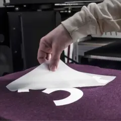Main tenant une feuille de transfert pour flocage fer à repasser sur un t-shirt violet.