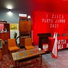 Scena imprezy w stylu PRL: meble, radio, telewizor, akordeon, czerwony transparent z napisem.