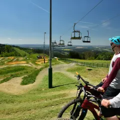 Zwei Mountainbiker mit Helmen blicken auf einen aufregenden mountainbike trail mit Sprüngen und Wellen. Eine Seilbahn schwebt darüber.