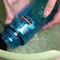Hände spülen eine blaue Nalgene-Flasche mit kochendem Wasser in einer grünen Schüssel.