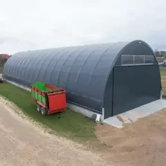 Duża, szara hala namiotowa z zielono-czerwonym przyczepą rolniczą obok.