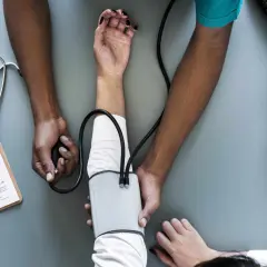 Lekarz mierzy ciśnienie pacjentowi. Na stole leży stetoskop, dokumentacja medyczna i ciśnieniomierz, który pokazuje ciśnienie rozkurczowe.
