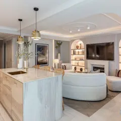 Elegante salón-comedor con isla de cocina. Una lámpara de 4 focos ilumina la zona de estar, destacando el sofá curvo y la chimenea.