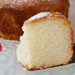 Una fetta di torta senza glutine, soffice e dorata, cosparsa di zucchero a velo, su un piatto trasparente.