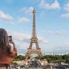 Kobieta robi zdjęcie telefonem ikonicznej wieży Eiffel. Paryski krajobraz z zielenią i budynkami w tle.
