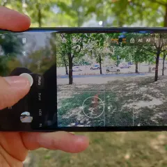 Dłoń trzyma smartfon, na ekranie którego widać interfejs aparatu. Aplikacja aparatu jest gotowa do zrobienia zdjęcia.