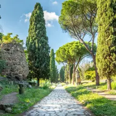 Allée bordée de cyprès et de pins sur la Via Appia, avec des ruines antiques et un ciel bleu.