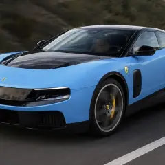 Una Ferrari elettrica blu cielo sfreccia su una strada, con il cofano nero e cerchi sportivi.