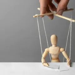 Drewniany ludzik-marionetka, sterowany przez ludzką dłoń, symbolizuje podżeganie i manipulację.