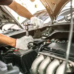 Mechanik w białych rękawiczkach dokręca śrubę kluczem nasadowym. Silnik samochodu jest otwarty, co sugeruje przegląd samochodu po terminie.