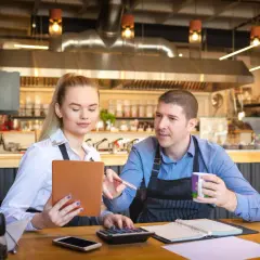Para omawia koszty uzyskania przychodu w restauracji, analizując dane na tablecie i kalkulatorze.