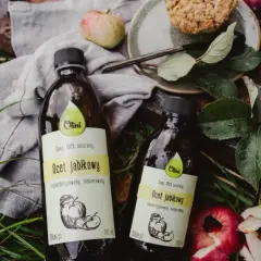 Dwie butelki naturalnego octu jabłkowego Olini, idealnego dla dzieci. Wokół jabłka i liście.