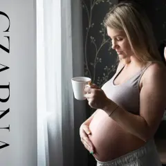 Ciężarna kobieta z kubkiem czarnej herbaty w dłoni, spogląda przez okno. Delikatne światło podkreśla jej brzuszek.