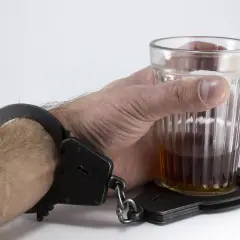 Ręka w kajdankach trzyma szklankę z alkoholem. Symbolizuje to problem, co zrobić z alkoholikiem awanturnikiem, który jest uwięziony w nałogu.