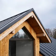 Tetto spiovente in metallo nero e legno naturale, con una finestra triangolare. Il legno, trattato per resistere agli agenti atmosferici, mostra il suo **impregnato significato** di protezione e calore.