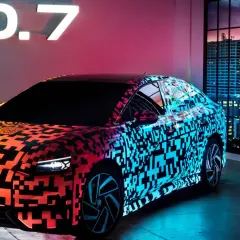 Nowy Volkswagen ID.7 prezentuje się w neonowym blasku, z efektownym wzorem QR kodu na karoserii. Obok stoi mężczyzna w garniturze, a w tle widać nocną panoramę miasta.