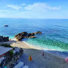 Albania, czerwiec: słoneczna pogoda, turkusowe morze, kamienista plaża z parasolami i łódkami na horyzoncie.