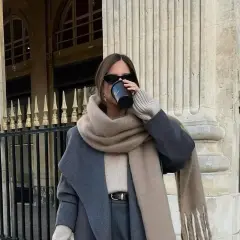 Stilvolle Frau trinkt Kaffee, kombiniert grau und beige mit einem Oversize-Schal und Mantel.