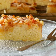 Ein saftiges Stück mandelkuchen vom blech, bestreut mit Mandelsplittern und Zucker, liegt auf einem Glas-Teller.